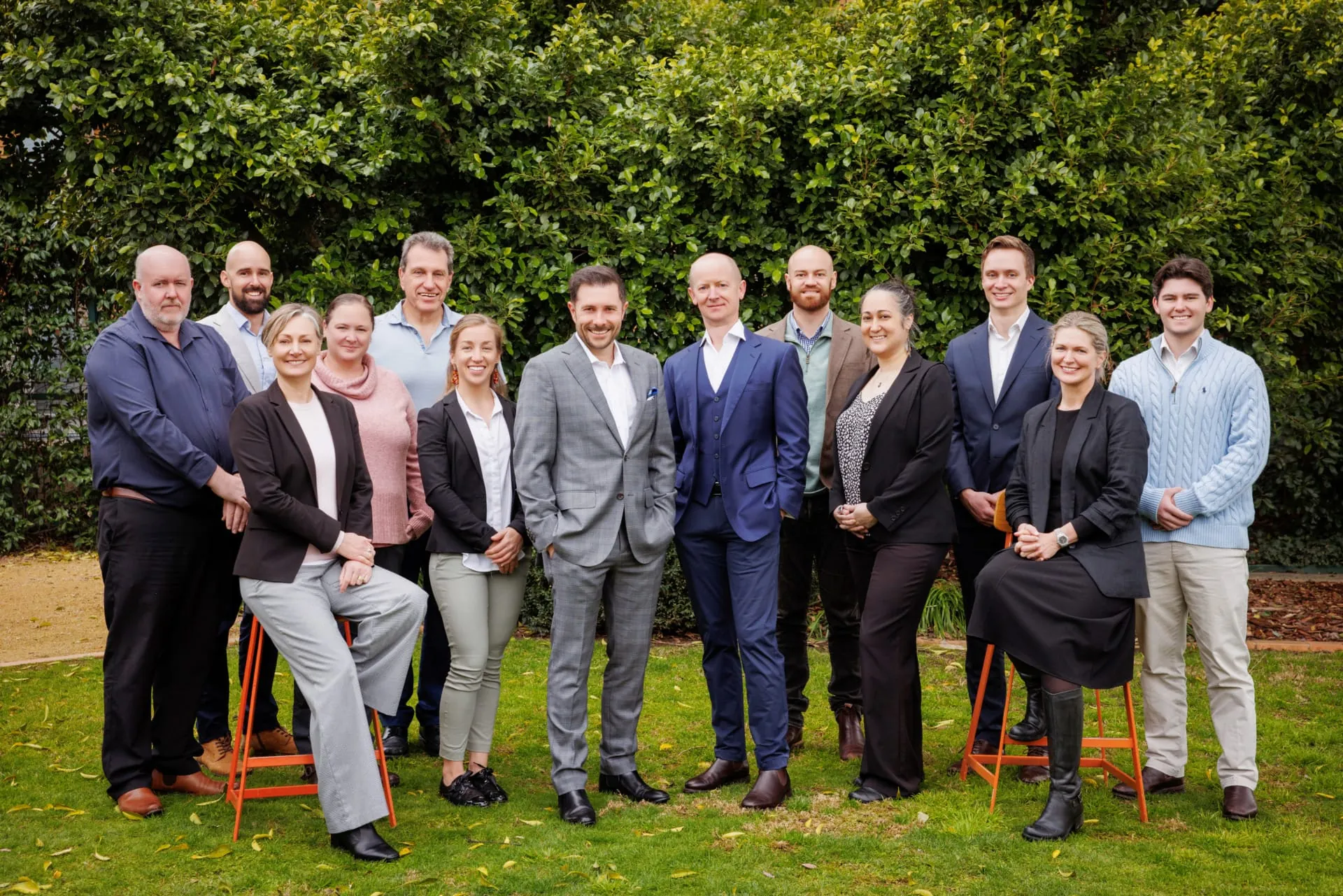 Thirteen professionally dressed colleagues, men and women, standing and sitting on orange stools on a grassy lawn and smiling toward the camera with a tall leafy hedge behind them.
